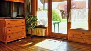 Interior em madeira com radiador, cómoda de madeira, planta em vaso e porta de correr de vidro que dá para área externa.