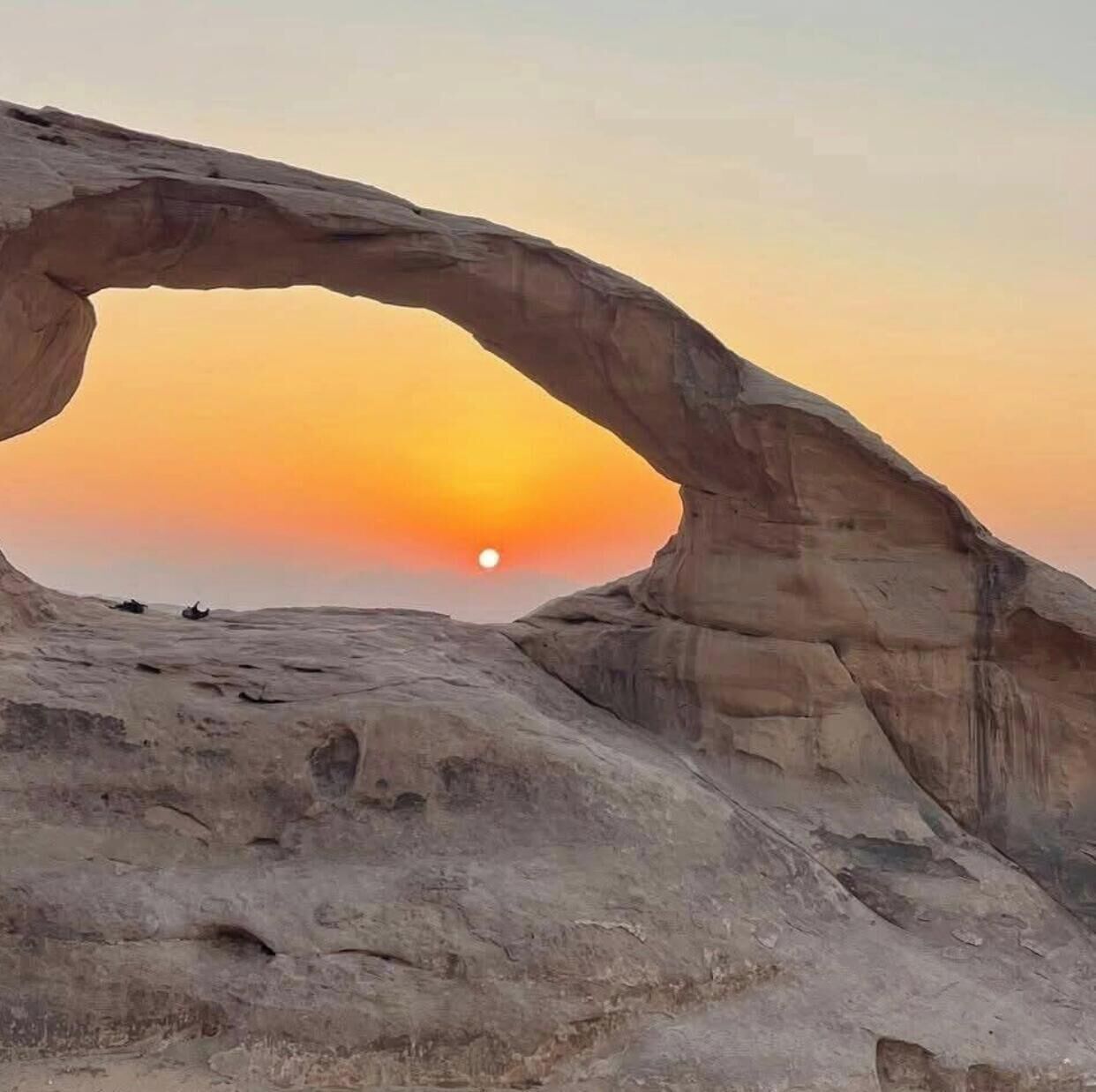 A natural rock formation arching over a flat landscape with a sunset in the background.