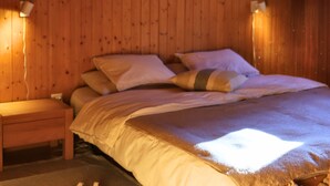 A wooden-paneled bedroom with a bed, pillows, a nightstand, and a pair of slippers on a mat.