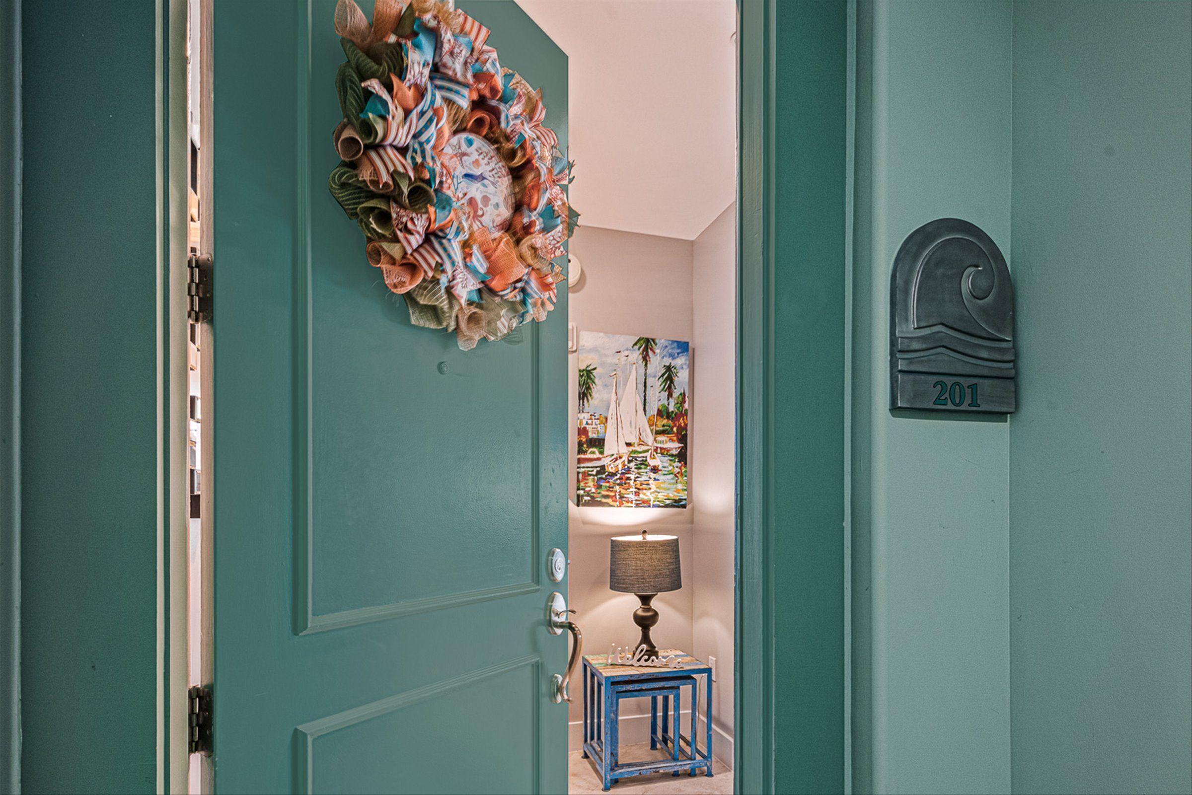 Una puerta color turquesa con un adorno festivo, una mesita con una lámpara y un cuadro de veleros en la pared.