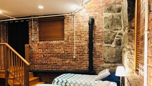 A room with a brick wall, a wooden staircase, a bed with patterned bedding, and a nightstand with a lamp. - Enjoyable 2-bedroom lower level apartment in charming Lynn (Lynn)