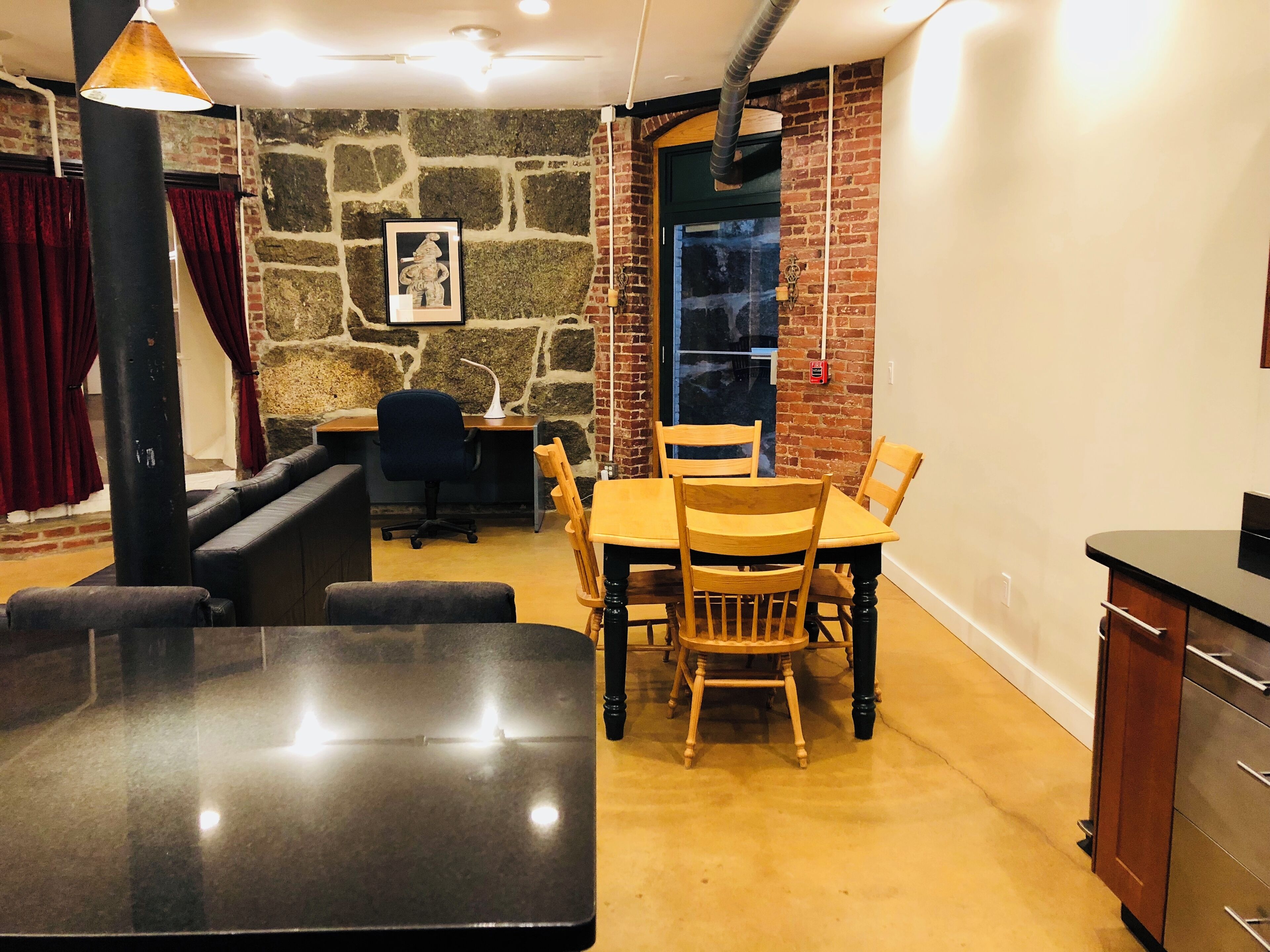 A cozy room with a stone wall, a wooden dining table with chairs, a black sofa, a desk with a chair, and a pendant light.