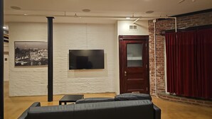 A living room with a black leather sofa, a flat-screen TV mounted on a white brick wall, a wooden door, and a red curtain. - Enjoyable 2-bedroom lower level apartment in charming Lynn (Lynn)