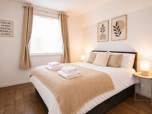 A neatly made bed with a beige blanket and pillows, a bedside lamp, and a window with curtains.