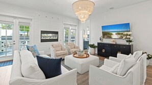 A spacious living room with a large chandelier, white sofas, and a view of the outdoors through large windows. - Golf Cart Perks, Large Private Pool, Close to Beach (Santa Rosa Beach)