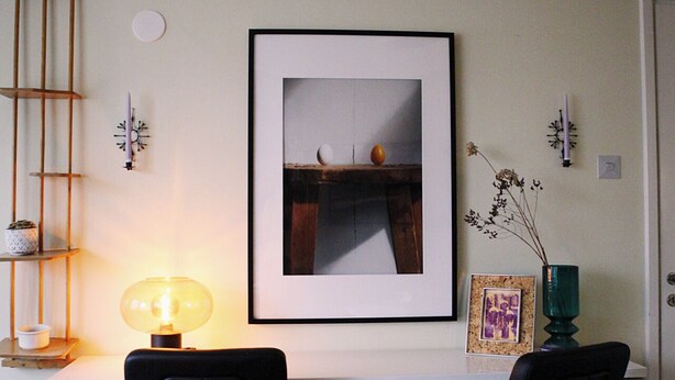 A room with a white desk, two black chairs, a framed picture on the wall, a small table with a lamp, and a vase with dried flowers.