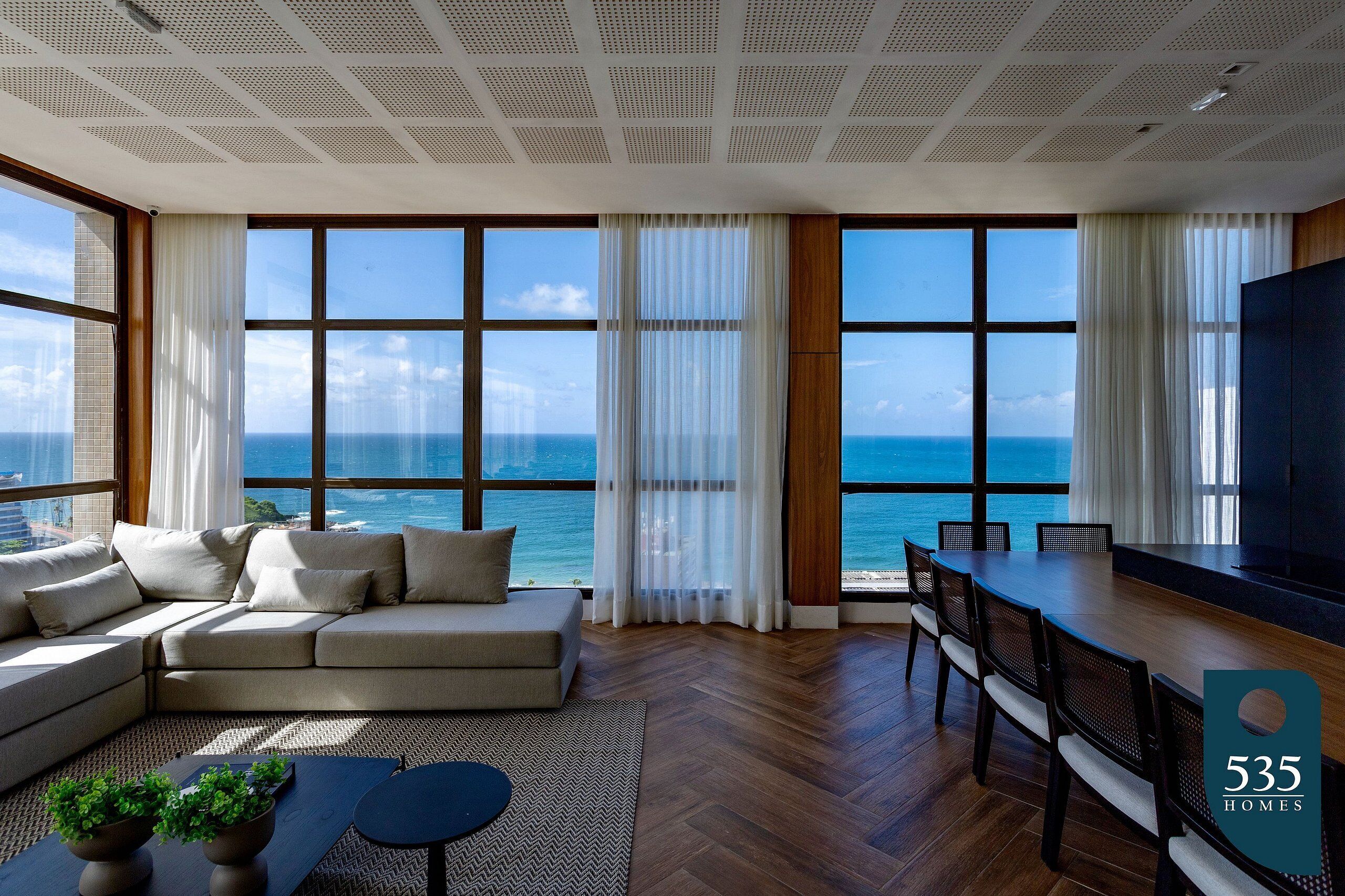 A modern living room with a large sofa, dining table, and chairs, large windows with ocean views, and a flat-screen TV.