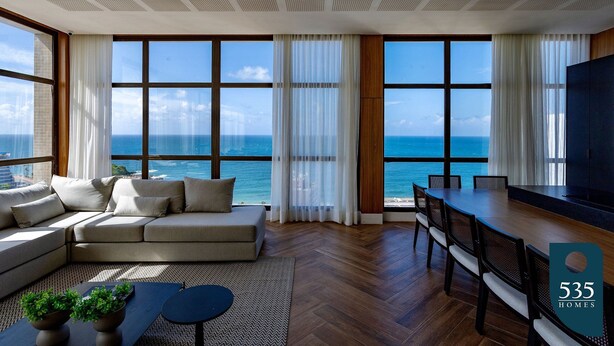 Moderni olohuone, jossa on suuri sohva, ruokapöytä ja tuolit, suuret ikkunat, joista avautuu näkymä merelle, sekä litteänäyttöinen televisio.