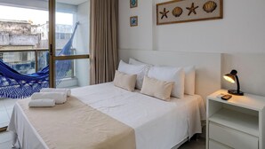 Ett hotellrum med en säng, ett nattduksbord, en lampa, utsikt över staden och en hängmatta utanför fönstret.