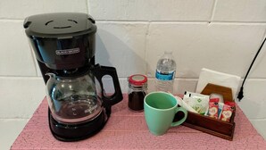 Una cafetera, una taza, una botella de agua, un tarro y una caja de cápsulas de café sobre una superficie con baldosas rosas.