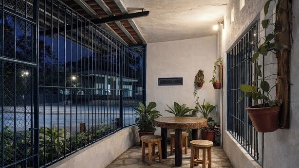 Un pequeño patio al aire libre con una mesa redonda de madera y taburetes, rodeado de macetas con plantas y una valla de metal.