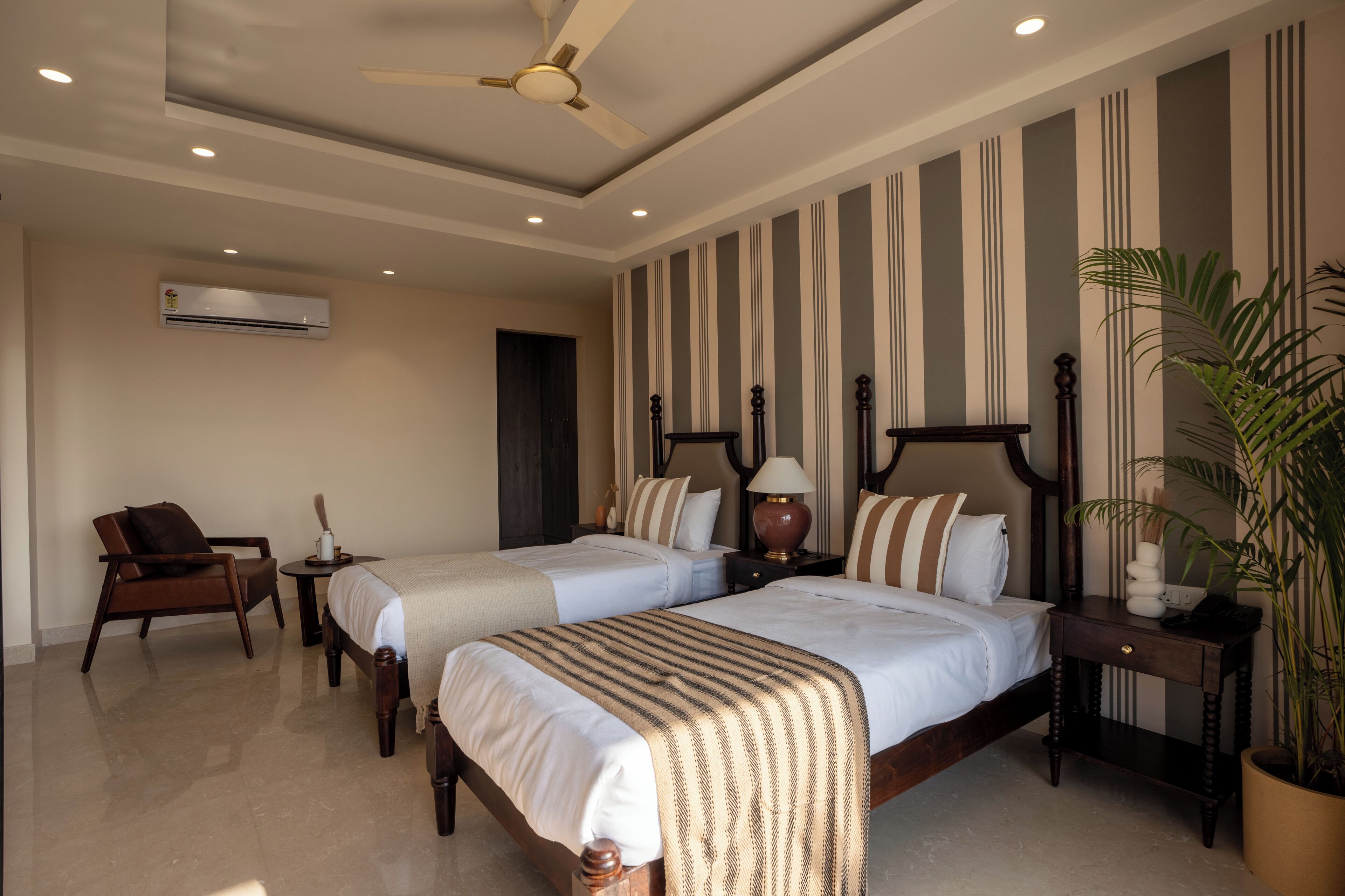 A hotel room with two beds, a ceiling fan, an air conditioning unit, a chair, a nightstand, and a potted plant.