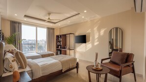A modern hotel room with a large bed, a seating area with a chair and a small table, a TV mounted on the wall, and a view of the cityscape through large windows. - The Hi-rise (Gurugram)