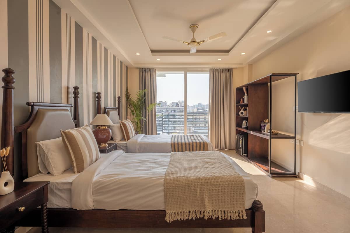 A bedroom with two beds, a ceiling fan, a large window with curtains, a bookshelf, and a flat-screen TV.