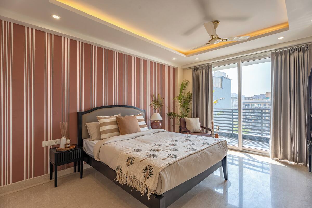 A modern bedroom with a large bed, a nightstand, a lamp, and a view of the city through large windows.