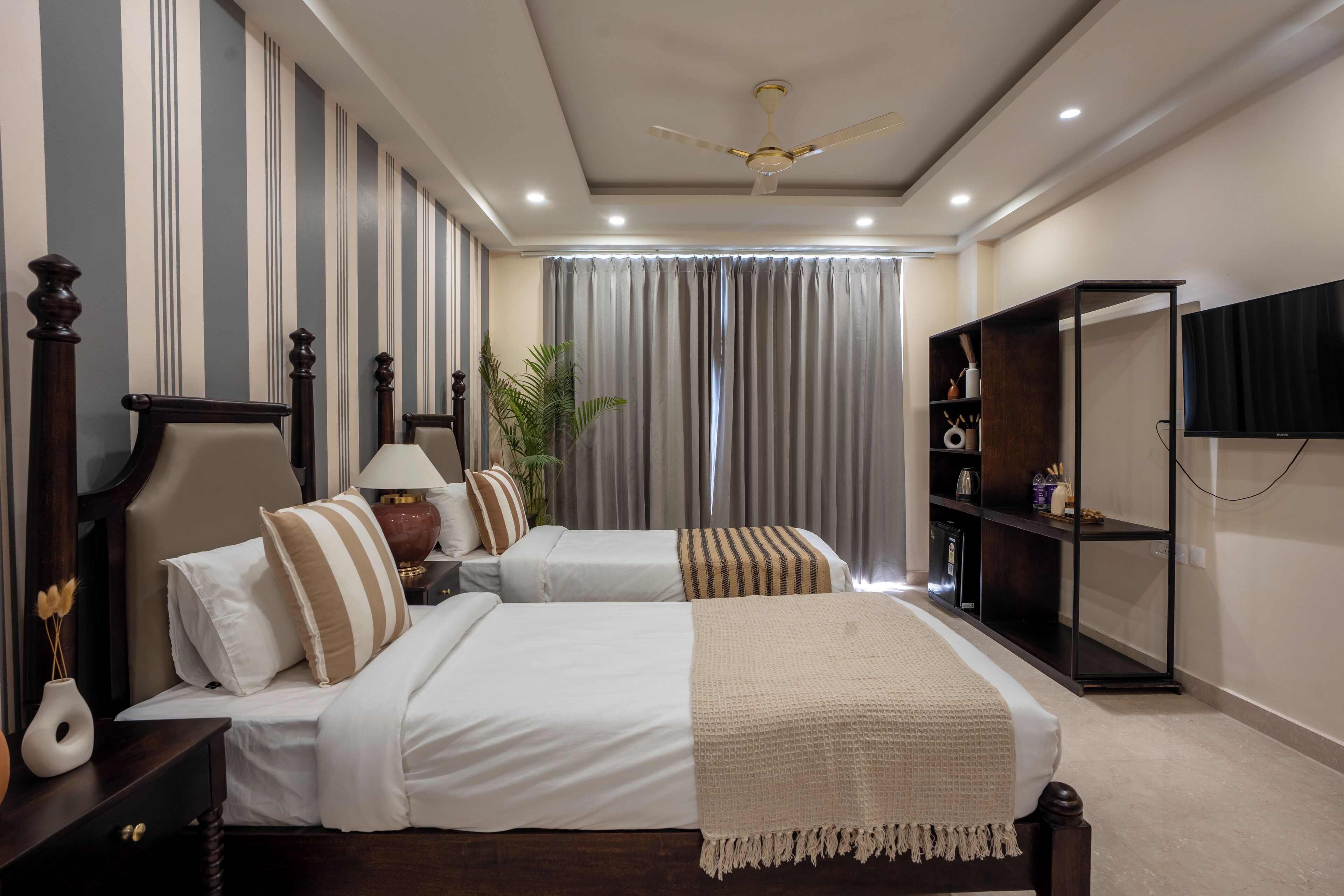 A hotel room with two beds, a ceiling fan, a TV, a bookshelf, and a large window with curtains.