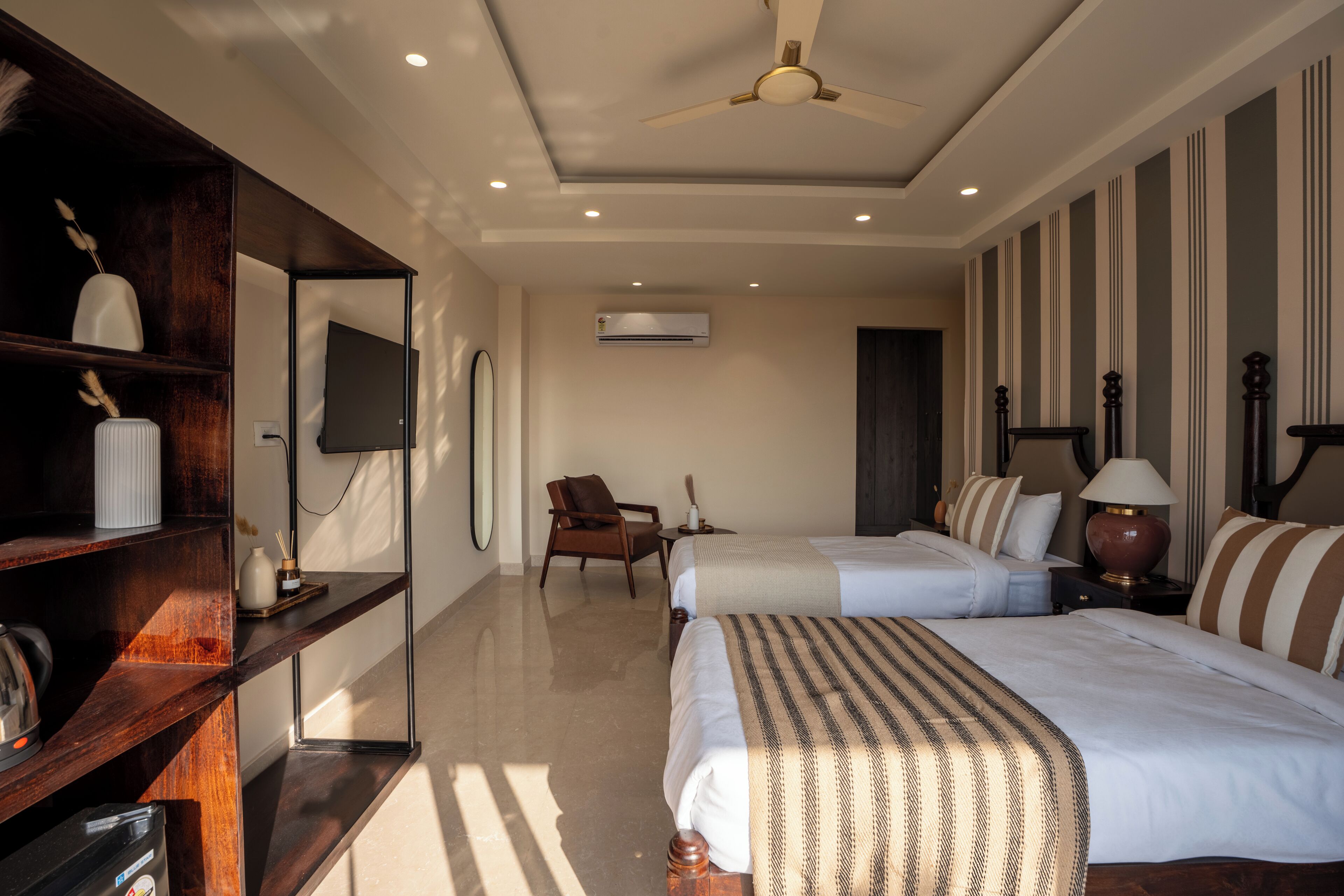 Une chambre d’hôtel avec deux lits, une étagère en bois ornée d’objets décoratifs, un téléviseur, un ventilateur de plafond et un appareil de climatisation.
