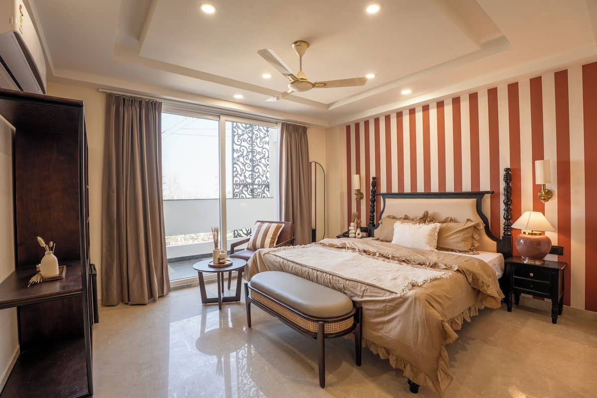 A bedroom with a large bed, a ceiling fan, a bedside table with a lamp, a bench, a small table with a vase, and a view of the outdoors through a window.