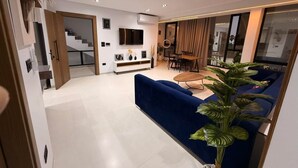 Ein modernes Wohnzimmer mit einem blauen Sofa, einem Couchtisch und einem Essbereich mit Tisch und Stühlen. Eine Treppe führt zu einer weiteren Ebene, und ein großes Fenster mit Vorhängen.