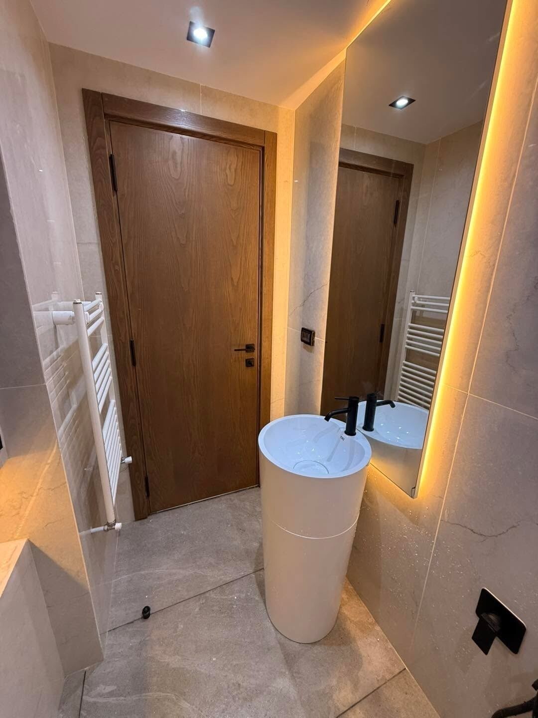 Uma casa de banho com porta de madeira, lavatório branco com torneira preta e um espelho a refletir o espaço.