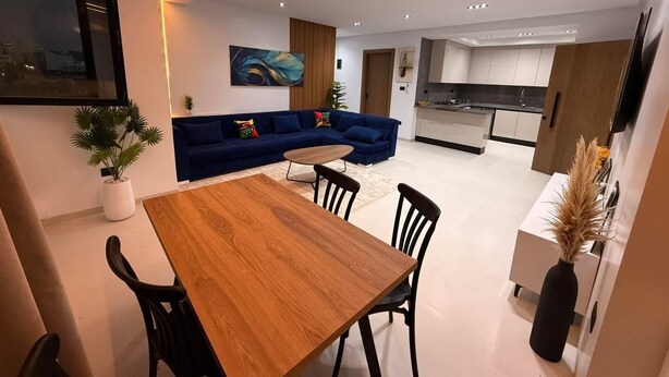 Ein modernes Wohnzimmer mit einem Holzesstisch, schwarzen Stühlen, einem blauen Sofa und einer Küche im Hintergrund.