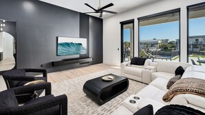 A modern living room with a flat-screen TV, black and white furniture, a ceiling fan, and large windows offering a city view. - Isle Be Back|Private Pool|Waterfront|Golf Cart (Destin)