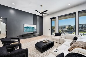 A modern living room with a flat-screen TV, black and white furniture, a ceiling fan, and large windows offering a city view. - Isle Be Back|Private Pool|Waterfront|Golf Cart (Destin)