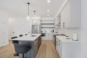Cozinha moderna com armários brancos, uma ilha central com banquetas, eletrodomésticos de aço inoxidável e iluminação pendente.