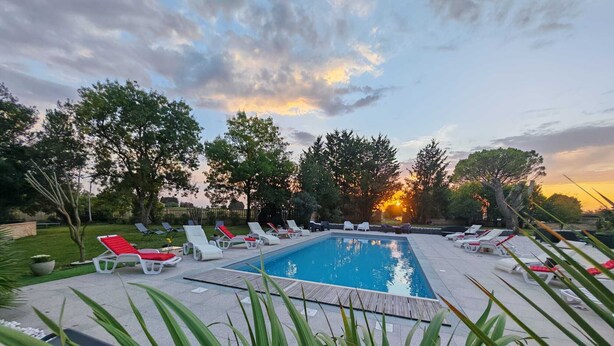 Ein rechteckiger Swimmingpool, umgeben von Liegestühlen mit roten Kissen, eingebettet in einen angelegten Garten mit Bäumen und einem Sonnenuntergang im Hintergrund.