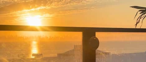 A sunset view from a balcony with a railing, overlooking a cityscape and a body of water.