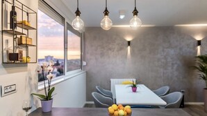 Un espace repas moderne comprenant une table, des chaises, un saladier de fruits, une plante en pot et une vue sur la ville par la fenêtre.
