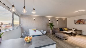 Un salon moderne avec un canapé gris, une table à manger avec des chaises et un coin cuisine agrémenté d’un panier de fruits.