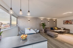 Ein modernes Wohnzimmer mit einem grauen Sofa, einem Esstisch mit Stühlen und einer Küchenzeile mit Obstschale.