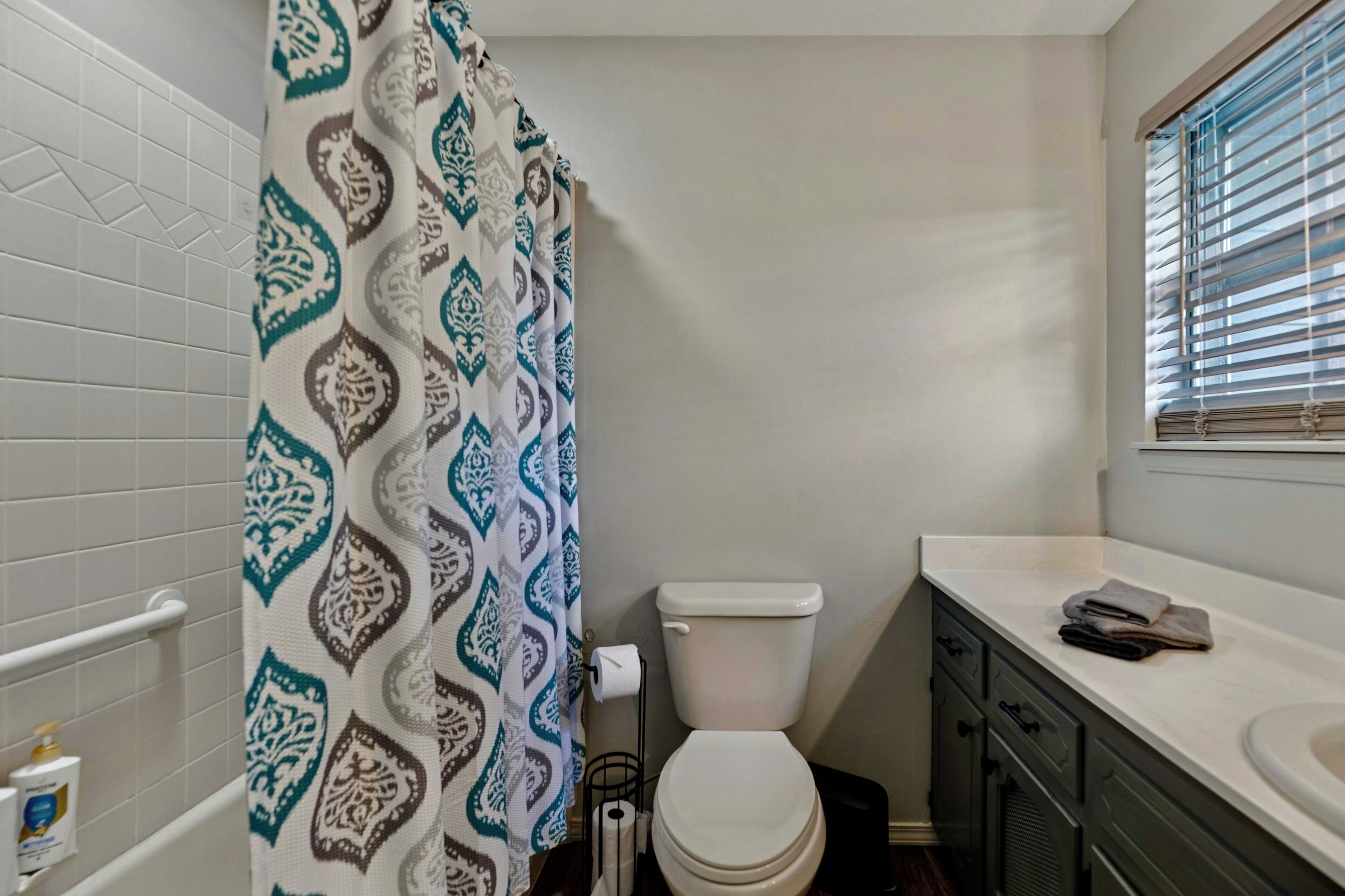 A bathroom with a patterned shower curtain, a toilet, a sink, and a window with blinds.