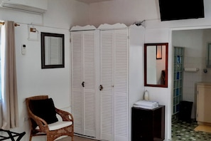 A room with a wicker chair, a white wardrobe, a small table with a white towel, a mirror, and a bathroom visible through an open door. - Hillside Penthouse large 2-bed/bath condo, retro feel, modern appliances (Dutch Cul de Sac)