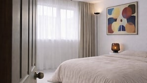 A bedroom with a large bed, a hanging light fixture, a painting on the wall, and a door with a round doorknob.