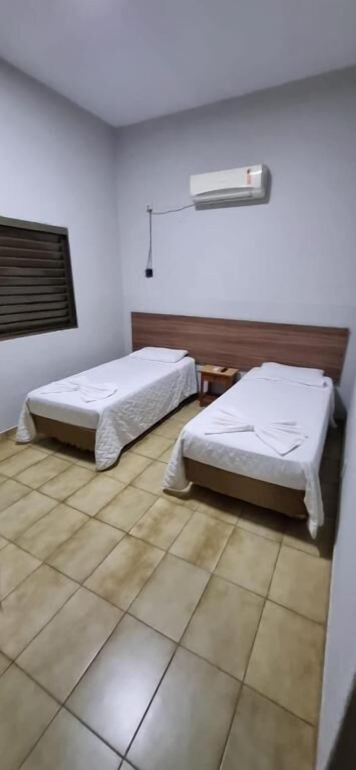 A room with two single beds, a wooden headboard, a window with blinds, and a wall-mounted air conditioner.