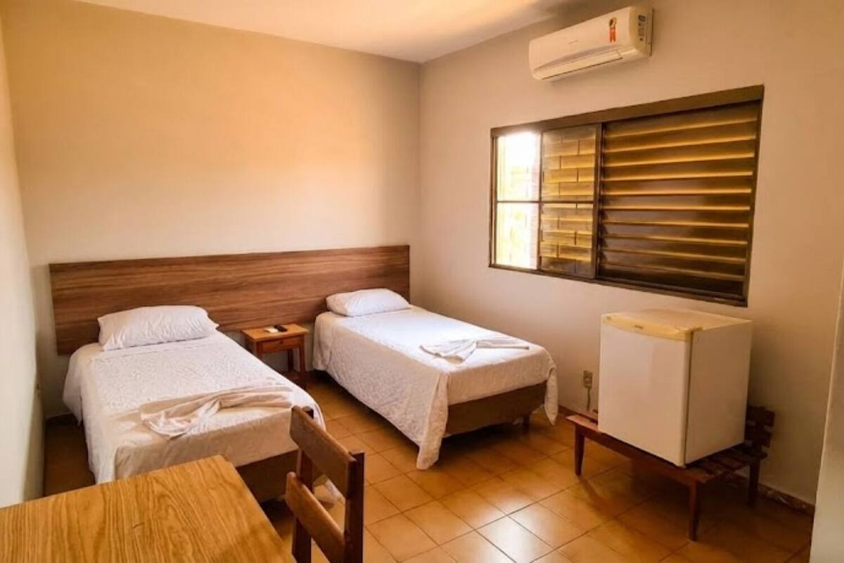 Una habitación pequeña y funcional con dos camas individuales, una mesa de madera, una silla, un escritorio pequeño y un radiador.