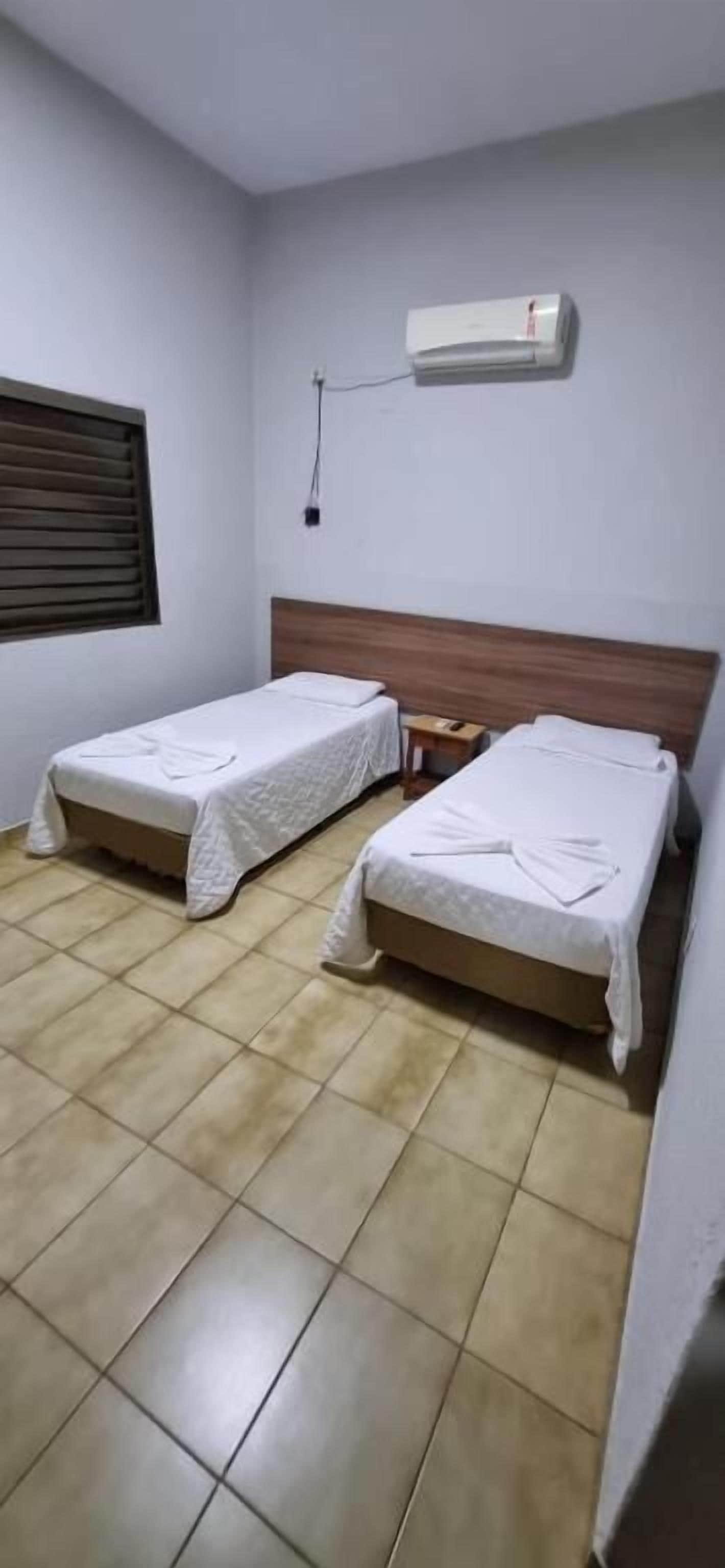 A room with two single beds, a wooden headboard, a wall-mounted air conditioner, and a window with blinds.
