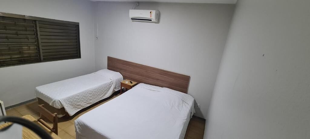 A small, clean room with two single beds, a wooden headboard, a small table, and a window with blinds.