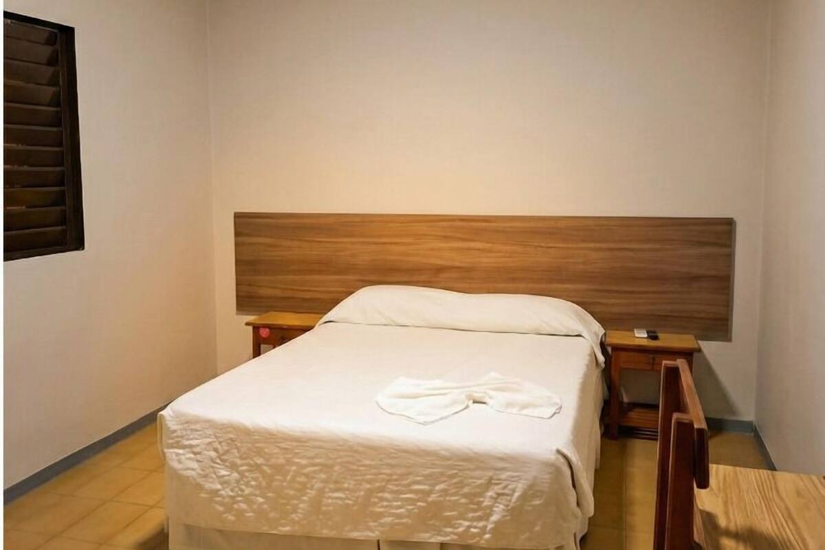 Un dormitorio con una cama individual, cabecera de madera, mesita de noche, una silla de madera y una pequeña mesa con un control remoto.