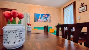 A dining area with a wooden table, chairs, a vase with red flowers, a painting on the wall, and a view of a cityscape through a window. - VILLA VENEZIA (Mira)