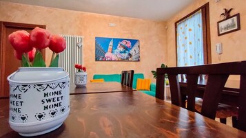 Un'area pranzo con un tavolo in legno, sedie, un vaso con fiori rossi, un quadro al muro e vista sulla città attraverso una finestra.
