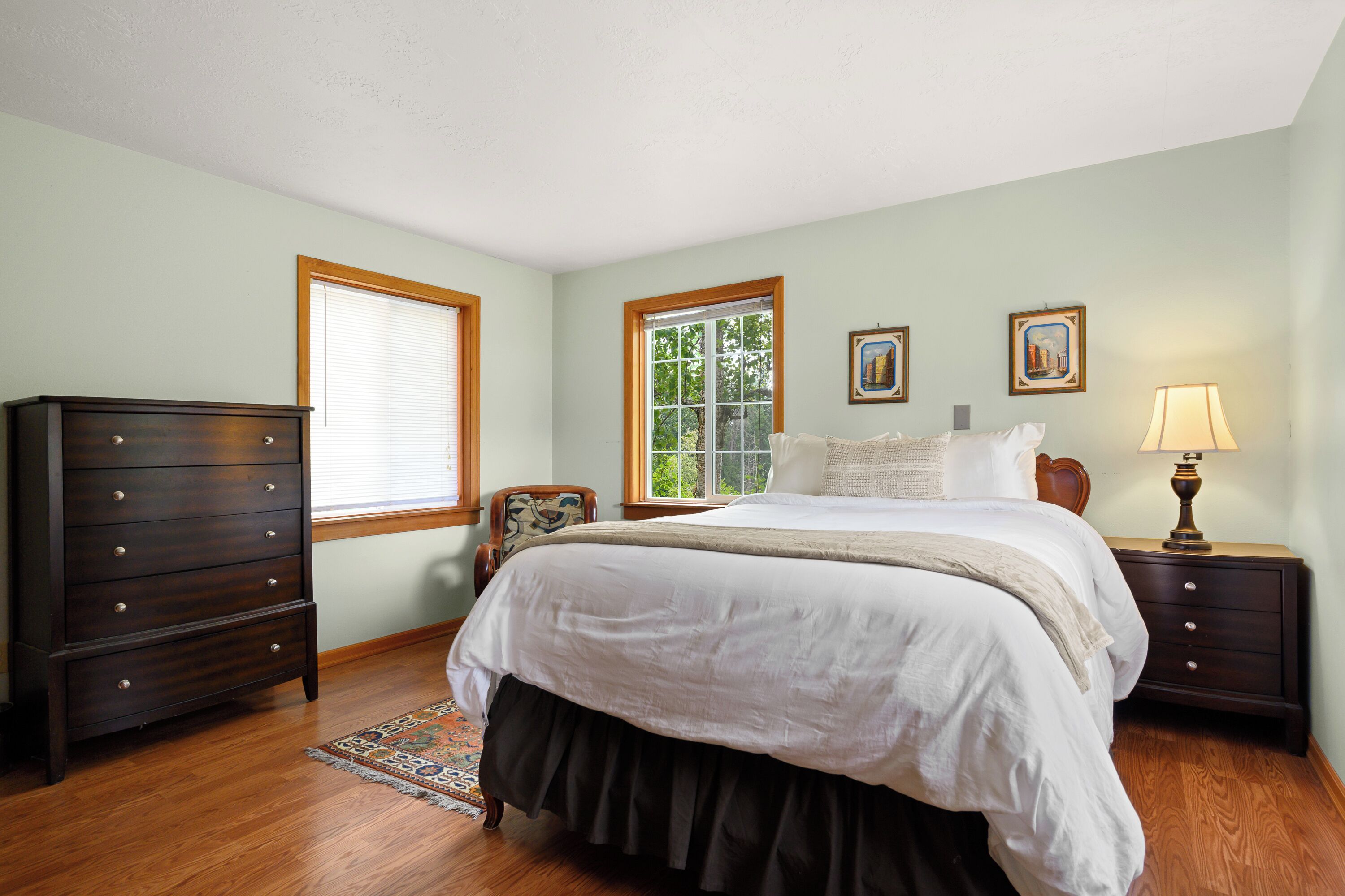 A bedroom with a bed, dresser, nightstands, lamp, and two windows.