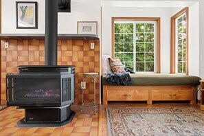 A cozy room with a wood-burning stove, a bench with cushions, and a large window with a view of greenery. - Waterfront Retreat with Private Dock & Lake Views (Sandpoint)