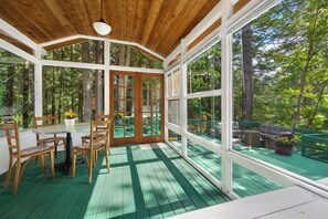 Un espacio exterior techado con una mesa y sillas de madera, ventanas amplias y vistas a un bosque.