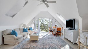 Geräumiges Schlafzimmer im Dachgeschoss mit hohen Decken, einem Deckenventilator, einem großen Fenster mit Blick auf Bäume, einem Bett mit Holzrahmen, einem Fernseher und einem Essbereich mit Tisch und Stühlen.