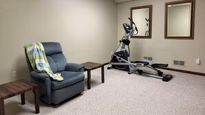 A room with a grey armchair, a standing exercise machine, a small table, and two mirrors on the wall.