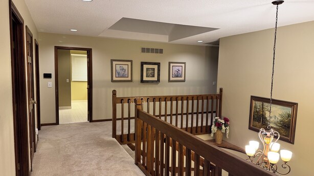 A hallway with a wooden railing, carpeted floor, and framed pictures on the wall.