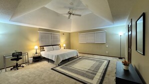 A bedroom with a bed, two bedside tables, a desk with a chair, a standing lamp, a ceiling fan, and a large window with blinds.